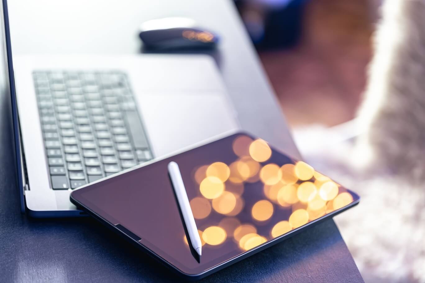 Tablet and stylus on a desk beside a laptop with bokeh lights